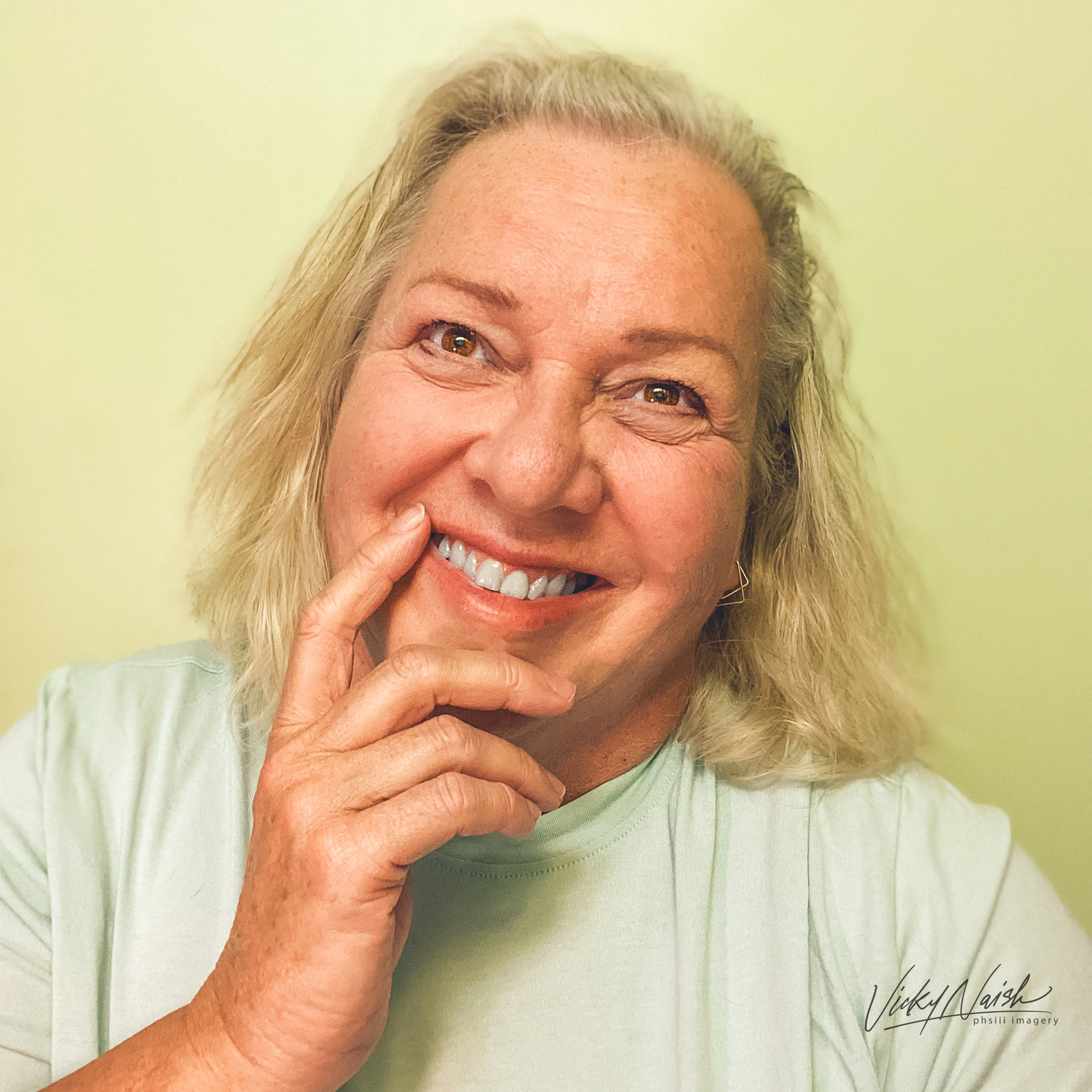 portrait of smiling blonde woman in pale green shirt on pale yellow background  woman has hand gently place by her chin and mouth and is looking forward with her eyes looking up